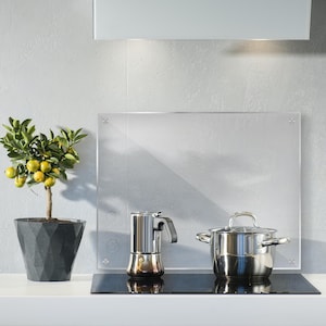 May include: A stainless steel pot and a silver coffee maker sit on a black electric hob with a glass splashback. A small potted citrus tree with green leaves and yellow fruit sits on a white worktop.