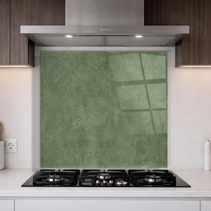 Puede incluir: Una cocina moderna con una encimera negra y un protector contra salpicaduras verde. El protector contra salpicaduras tiene una apariencia texturizada, similar al mármol. Los armarios de madera oscura están encima de la encimera y una campana extractora de acero inoxidable está instalada encima.