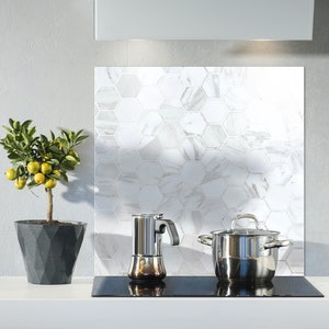 May include: A white kitchen backsplash with a hexagonal tile pattern. A small potted citrus tree sits on the counter next to a stovetop with a silver pot and a silver coffee maker.