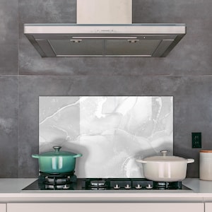 Puede incluir: Una escena de cocina moderna con una campana extractora de acero inoxidable sobre una placa de cocina con dos ollas. Una olla verde turquesa está en el quemador izquierdo, y una olla color crema está a la derecha. Una tetera y botes blancos están en la encimera.