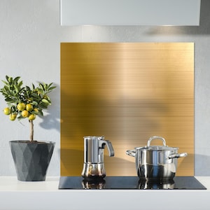 May include: A gold metal backsplash with a brushed finish, a black electric stovetop with a pot and a coffee maker on it, and a small potted lemon tree to the left.