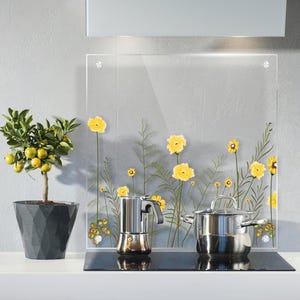 Puede incluir: Una placa protectora de cocina de vidrio transparente con un diseño floral de flores amarillas y tallos verdes. Una olla y una cafetera de acero inoxidable están sobre una placa de cocina negra. Un árbol de cítricos en maceta está en la encimera.