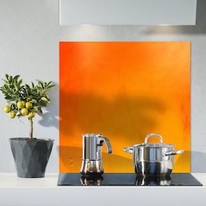 Op de afbeelding: Een roestvrijstalen kookplaat met een zwarte glazen kookzone. Een zilveren pan en een zilveren koffiezetapparaat staan op de kookplaat. De achterwand is oranje en geel.