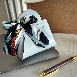 May include: A light blue leather handbag with a top handle and a patterned scarf tied around the handle. The bag is sitting on a laptop computer.