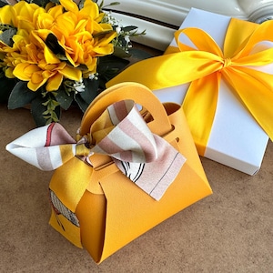 Könnte beinhalten: Eine kleine gelbe Handtasche mit einem gestreiften Schal, der um den Griff gebunden ist. Die Handtasche liegt auf einer braunen Oberfläche neben einer weißen Geschenkbox mit einem gelben Band.