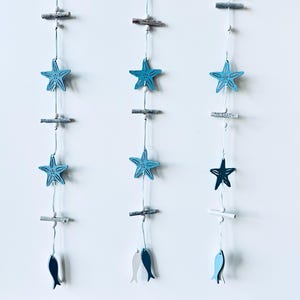May include: A coastal-themed wall decoration with three hanging strands. Each strand features blue starfish, wooden beads, and small fish cutouts in shades of blue and grey. The elements are suspended by thin, light-coloured string.
