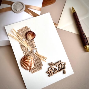 May include: A handcrafted birthday card with a seaside theme. The card is decorated with two seashells, faux pearls, and dried grass on a burlap patch. A wooden cutout spells "Happy Birthday". A pen and envelope are in the background.