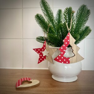 May include: Three wooden Christmas ornaments in red and white with white polka dots. A star, a tree, and a heart. The ornaments are hanging from a green pine branch in a white ceramic pot.