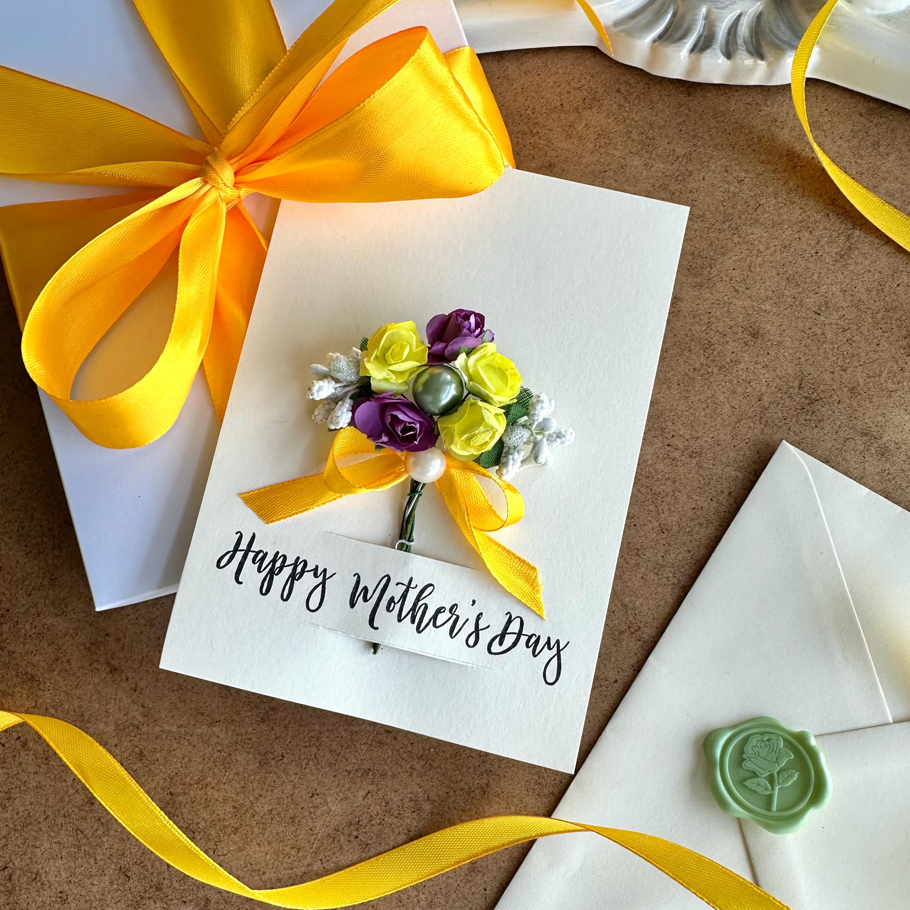 May include: A white card with a yellow ribbon and a bouquet of purple and yellow flowers with a pearl accent. The card reads "Happy Mother's Day".