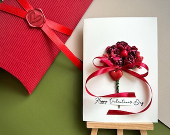 May include: A Valentine's Day card with a bouquet of red flowers and a red ribbon. The card is on a small wooden easel. A red gift box with a heart-shaped seal is in the background. The card says "Happy Valentine's Day."