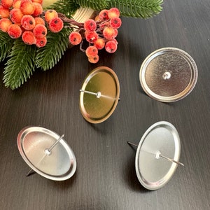 May include: Four silver and gold candle holders with a pin in the center. The candle holders are oval-shaped and have a metallic finish. The background includes a sprig of evergreen and red berries.