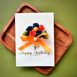 May include: A white rectangular greeting card with the text Happy Birthday in gold script. The card features a decorative bouquet of small dark blue and orange fabric roses, green accents, a peach-coloured bead, and an orange ribbon bow.