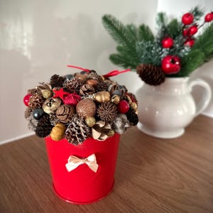 May include: A red metal bucket decorated with a small beige bow, filled with a dense arrangement of brown pinecones, gold glitter spheres, red berries, and a red star. The bucket sits near a white pitcher containing green pine branches and red berries.