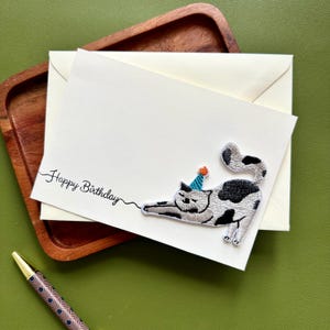 May include: A cream-coloured birthday card featuring a black and white cat wearing a party hat. The card has the script text "Happy Birthday". The card rests on a wooden tray, with a pen visible.