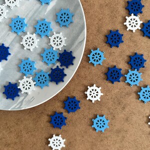 May include: A collection of blue, white and light blue wooden nautical wheel charms. The charms are scattered on a brown surface and a white plate.