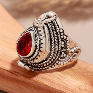 May include: A silver-colored ring with an ornate design. The ring features a teardrop-shaped, deep red gemstone. The band has detailed patterns, including braided and dotted textures. The ring is set on a wooden surface.