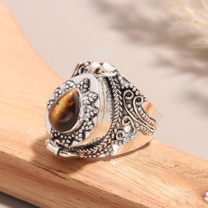 May include: A silver ring with an ornate design and a tiger's eye stone. The ring has a hinged lid that opens to reveal a small compartment.