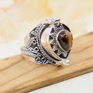 May include: A silver ring with an oval, brown gemstone. The ring has an ornate design with detailed scrollwork and a decorative setting around the stone. The ring is set on a wooden surface.