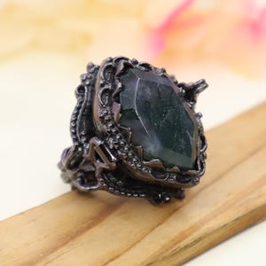 Puede incluir: Un anillo negro con una gran piedra preciosa verde oscuro engastada en un diseño detallado y ornamentado.