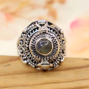 May include: A silver ring with an ornate design. The ring features a central, round stone with a gray-green hue, surrounded by detailed filigree and decorative elements. The ring is set on a wooden surface.