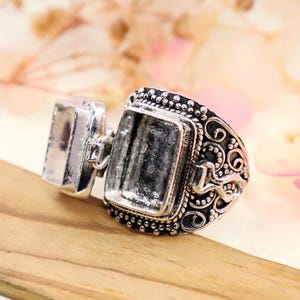 Peut inclure: Une bague argentée avec deux montures rectangulaires. La monture la plus grande présente un motif orné et décoratif sur les bords. La bague est présentée sur une surface en bois avec un arrière-plan floral flou.