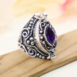 May include: A silver ring with an ornate design and a purple amethyst stone. The ring is a hinged box that opens to reveal a small compartment.
