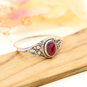 May include: A silver ring featuring a deep red oval gemstone. The ring has an intricate design with filigree detailing around the gemstone and on the shoulders of the band. The ring is displayed on a wooden surface.