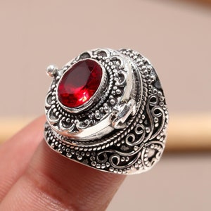 May include: A silver ring with an oval, deep red gemstone at the center. The ring has an ornate, detailed design with swirling patterns and a textured surface. The gemstone is set in a raised bezel.