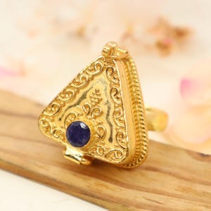 Może przedstawiać: Złoty pierścionek w kształcie trójkąta z klapką i małym niebieskim kamieniem szlachetnym osadzonym w środku. Pierścionek jest bogato zdobiony wzorem kwiatowym.