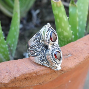 May include: A silver ring with two red gemstone accents. The ring has intricate floral designs and a hinged opening.