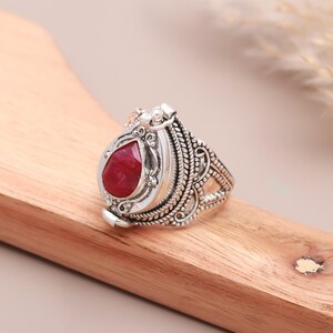 May include: A silver ring with an ornate design and a red teardrop-shaped stone. The ring is openable and has a hinged lid.