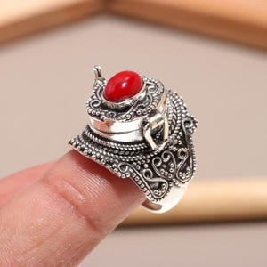 May include: A silver ring with an ornate design, featuring a small, oval, red stone at the top. The ring has intricate detailing around the band and setting, with a hinged top. The ring is on a finger.