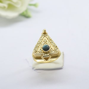 Puede incluir: Un anillo dorado con un colgante triangular. El colgante presenta intrincados diseños de filigrana y una pequeña gema oscura en el centro. El anillo está sobre una superficie blanca.