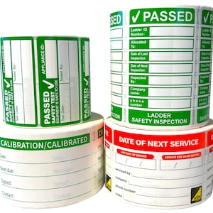 Op de afbeelding: Verschillende rollen witte en groene veiligheidsinspectielabels. De labels bevatten de tekst "PASSED", "SAFETY TEST" en velden voor informatie. Eén label luidt "LADDER SAFETY INSPECTION".