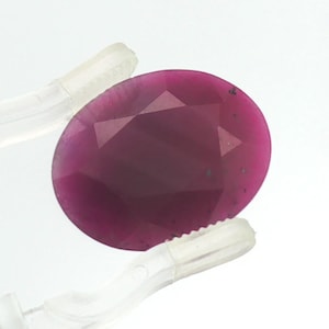 May include: An oval-shaped, deep magenta-coloured gemstone with a faceted surface. The stone is held by a pair of clear, translucent plastic tweezers, showcasing its translucent quality and the geometric cut of its top surface against a white background.