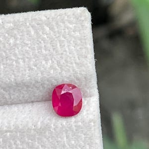 Puede incluir: Una gema de rubí de corte cojín vibrante con un color rojo intenso y una superficie facetada, exhibida en un soporte de exhibición texturizado de color claro. La gema muestra reflejos internos claros y un acabado pulido brillante.