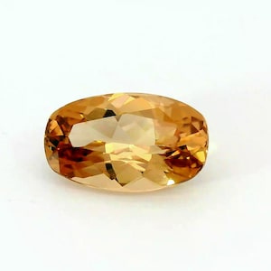 May include: An oval-cut, golden-yellow gemstone with facets. The gem has a polished surface that reflects light, creating a sparkling effect. The gemstone is set against a white background.