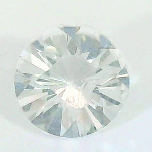 May include: A round, clear gemstone with multiple facets, reflecting light. The gemstone is set against a white background, showcasing its brilliance and clarity. The cut of the stone creates a starburst effect.