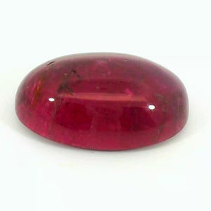 May include: A polished, oval-shaped gemstone with a deep red hue. The gem has a smooth, glossy surface with subtle internal inclusions and a slight translucence. The stone is set against a white background.