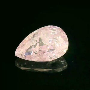 May include: A pear-shaped, light pink gemstone with a faceted surface. The gem is set against a black background, with its reflection visible below. The stone has a translucent quality, with internal inclusions.