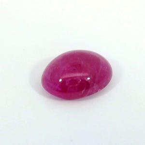 Pode incluir: Uma pedra preciosa de rubi polida, em forma oval. A gema é de uma cor vermelho-rosada profunda e saturada, com uma superfície lisa e refletora. A pedra está sobre um fundo branco liso.