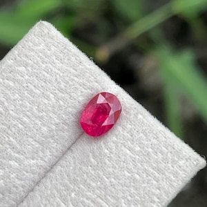 Puede incluir: Una gema roja vibrante de corte ovalado con una superficie facetada que refleja la luz. La piedra está colocada sobre una superficie de exhibición texturizada de color claro. El color carmesí profundo es consistente en toda la gema.