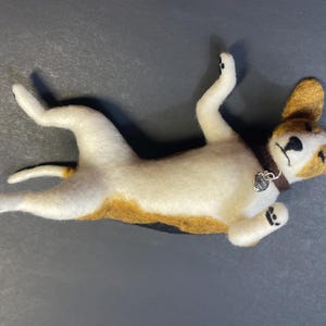 May include: A handmade felt dog figurine in a playful pose, lying on its back. The dog has a white body with brown and black markings, brown ears, and a brown collar with a small silver charm. The dog is approximately 6 inches long.