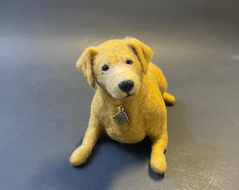 May include: A golden-yellow felted dog figurine, lying down with a small tag around its neck. The dog has a black nose and dark eyes, with a slightly textured appearance. The tag reads "Hand Made".