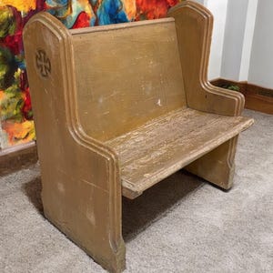 May include: A vintage wooden church pew with a distressed, aged appearance. The pew is painted in a light brown color with a cross carved into the side. The pew has a backrest and a seat, and is designed for one person.