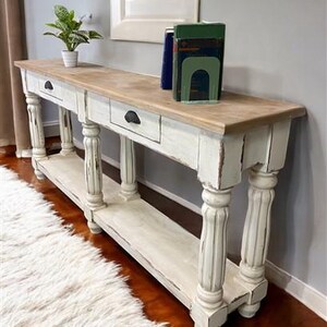 May include: White distressed console table with two drawers and a lower shelf. The table has turned legs and a rustic wood top.