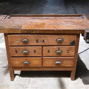 May include: A vintage wooden workbench with six drawers and a built-in vise. The drawers are labeled with letters and numbers. The workbench has a well-worn, aged appearance, with a rich, warm brown color. The top surface shows signs of use.