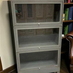 May include: A gray metal barrister bookcase featuring three stacked compartments, each with a clear glass lift-up door and a small round knob. The unit is supported by four metal caster wheels, allowing for mobility, and has a rectangular frame.