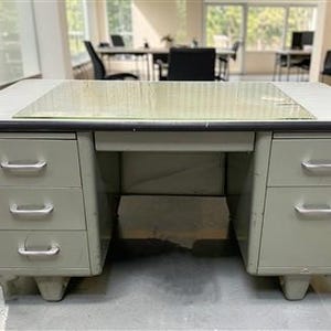 May include: A light gray metal office desk featuring a rectangular top with a clear glass protective cover, a central open knee space, and two pedestals. The left pedestal has three stacked drawers, while the right has two, all with silver handles.