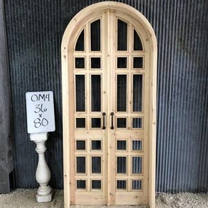 May include: A pair of arched wooden doors with a grid of glass panes. The doors are made of light wood and have black door handles. The doors are 36 inches wide and 80 inches tall.